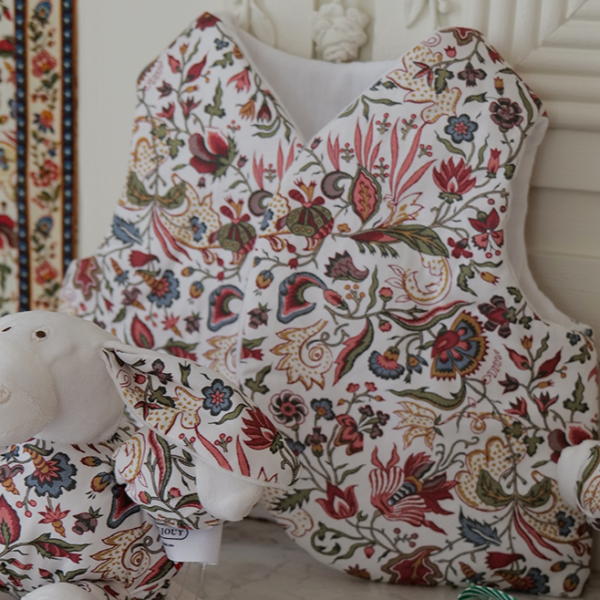 Un gilet matelassé à motifs floraux « FANTAISIE INDIENNE » de la marque BAVOIR est accroché à un panneau blanc, tandis qu’à l’avant, un mouton en peluche assorti est posé sur une surface en marbre — les deux pièces mettant en valeur le motif floral multicolore et finement travaillé de la collection. la marque Toile de Jouy