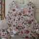 Un gilet matelassé à motifs floraux « FANTAISIE INDIENNE » de la marque BAVOIR est accroché à un panneau blanc, tandis qu’à l’avant, un mouton en peluche assorti est posé sur une surface en marbre — les deux pièces mettant en valeur le motif floral multicolore et finement travaillé de la collection. la marque Toile de Jouy