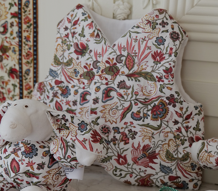 Un gilet matelassé à motifs floraux « FANTAISIE INDIENNE » de la marque BAVOIR est accroché à un panneau blanc, tandis qu’à l’avant, un mouton en peluche assorti est posé sur une surface en marbre — les deux pièces mettant en valeur le motif floral multicolore et finement travaillé de la collection. la marque Toile de Jouy