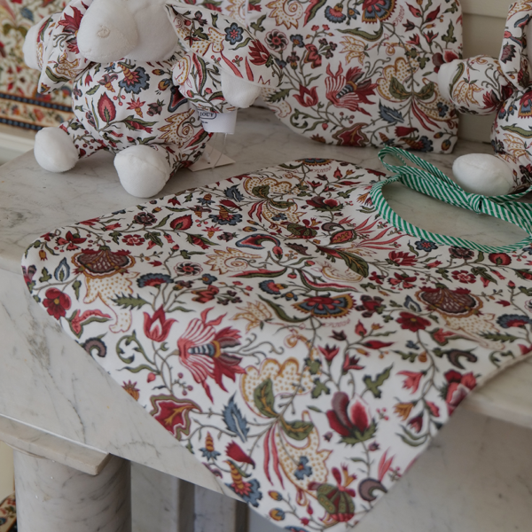Deux peluches et le bavoir « FANTAISIE INDIENNE » de BAVOIR sont posés sur un plan de travail en marbre. Tous ces articles arborent des motifs floraux colorés et raffinés, assortis les uns aux autres, sur fond blanc. la marque Toile de Jouy