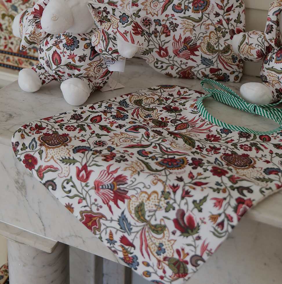 Deux peluches et le bavoir « FANTAISIE INDIENNE » de BAVOIR sont posés sur un plan de travail en marbre. Tous ces articles arborent des motifs floraux colorés et raffinés, assortis les uns aux autres, sur fond blanc. la marque Toile de Jouy
