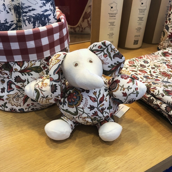 Le XCOMM_ELEPHANT BOULE FANTAISIE INDIENNE, un petit éléphant en peluche au corps blanc et aux oreilles, bras et jambes ornés de motifs floraux, est présenté sur un socle en bois, entouré de tissu et d'objets décoratifs. la marque Toile de Jouy