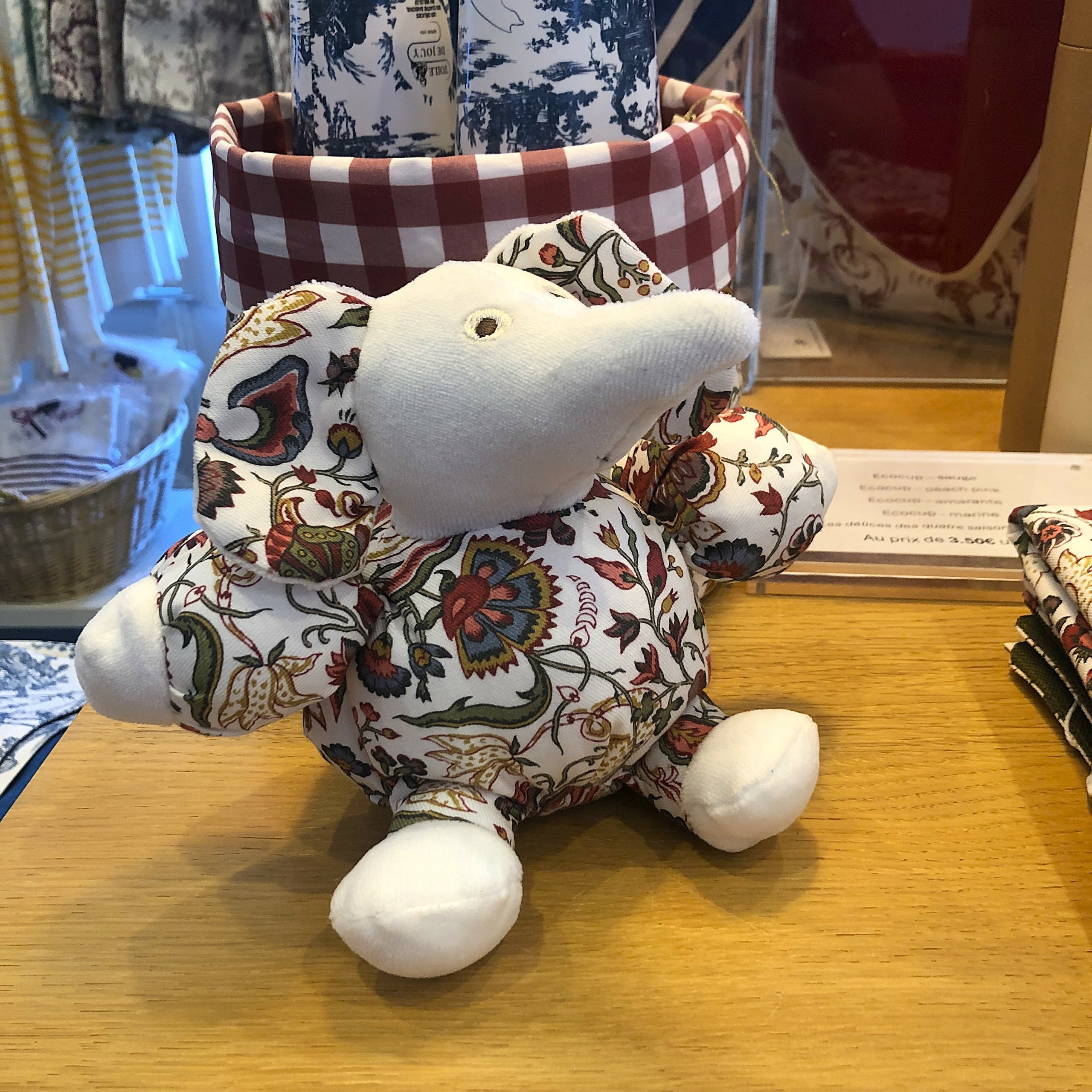 Le XCOMM_ELEPHANT BOULE FANTAISIE INDIENNE, une petite peluche en forme d'éléphant ornée d'un motif floral sur le corps et rehaussée de touches de tissu blanc sur les pattes, les bras et la trompe, est posée sur une table en bois à côté de tissus à motifs et de paniers. la marque Toile de Jouy