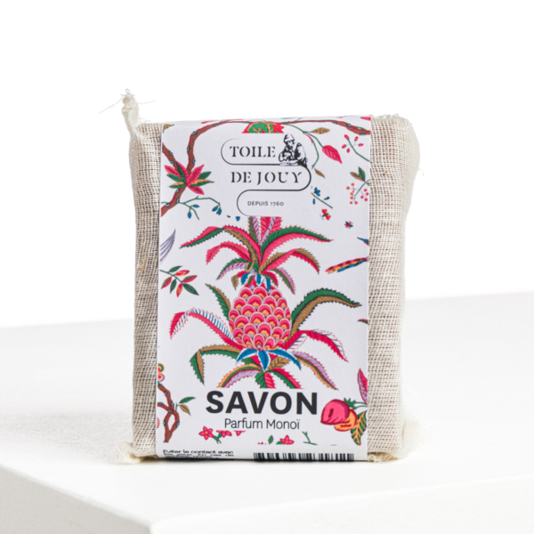 Une barre de XCOMM_SAVON ANANAS SENTEUR MONOÏ, enveloppée dans un filet blanc avec une étiquette vibrante d'ananas et de fleurs, repose sur une surface blanche. la marque Toile de Jouy