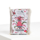 Une barre de XCOMM_SAVON ANANAS SENTEUR MONOÏ, enveloppée dans un filet blanc avec une étiquette vibrante d'ananas et de fleurs, repose sur une surface blanche. la marque Toile de Jouy