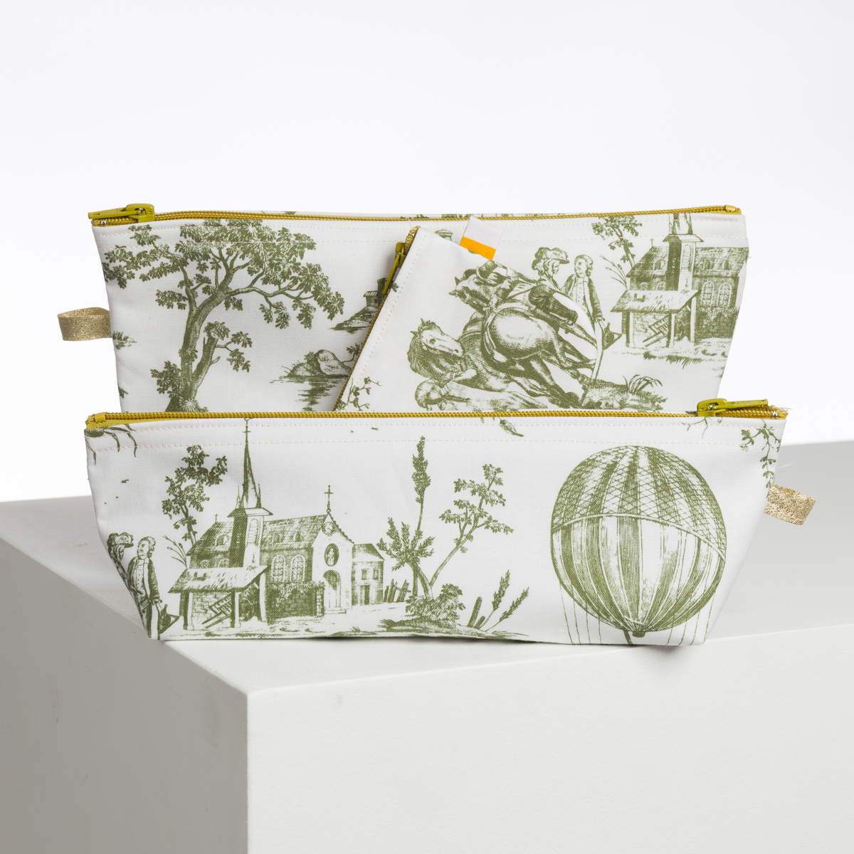 Le set XCOMM_BIC BALLON PESTO comprend deux pochettes en tissu à fermeture éclair jaune et des imprimés pastoraux verts - représentant une montgolfière, des arbres, des personnes et une église - représentés sur une surface blanche avec un fond blanc. la marque Toile de Jouy