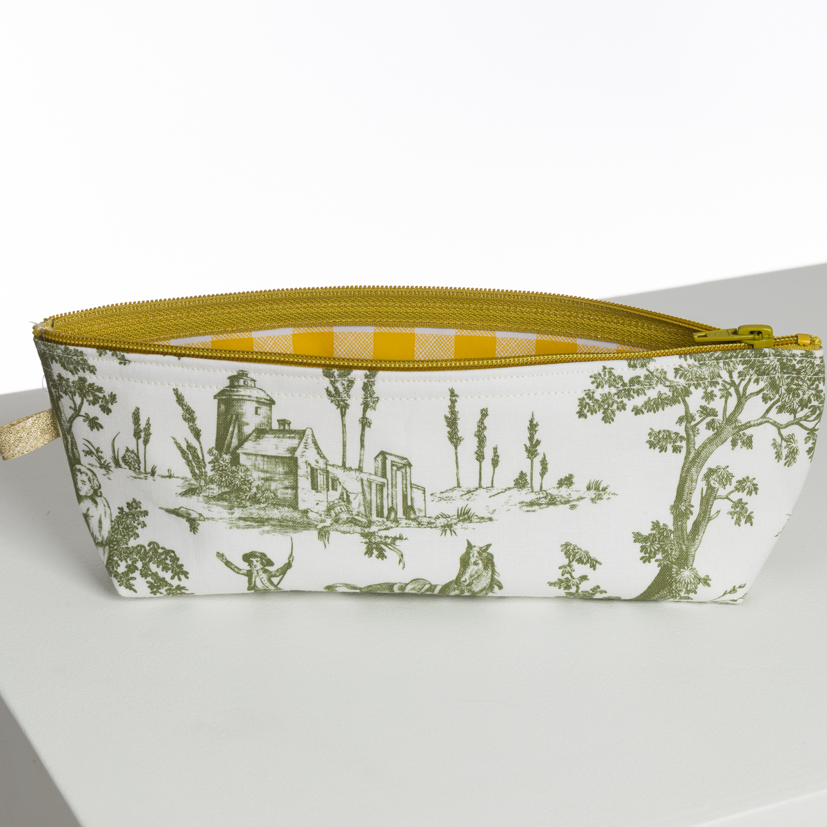 Le XCOMM_BIC BALLON PESTO est une pochette blanche à fermeture éclair avec des illustrations de toile pastorale verte et une fermeture éclair jaune. Présenté ouvert sur une surface blanche, son intérieur présente un motif à carreaux jaune et blanc. la marque Toile de Jouy