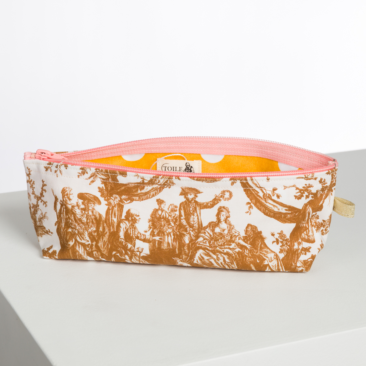Le XCOMM_BIC BALLON PESTO est une trousse rectangulaire avec une fermeture à glissière rose, une doublure à pois jaunes et un motif de toile marron et blanc de style vintage représentant des personnes en vêtements d'époque. Il est posé sur une surface gris clair. la marque Toile de Jouy