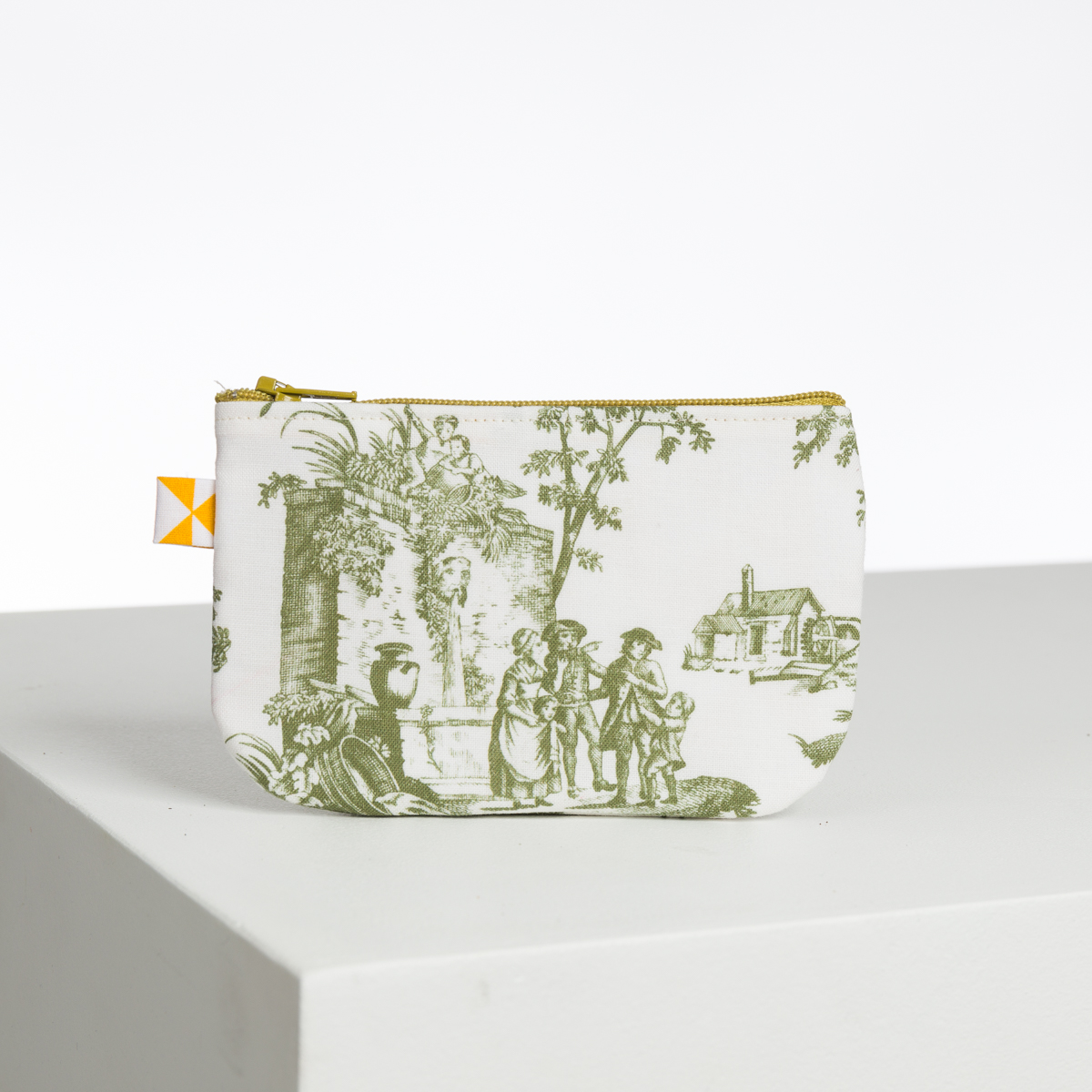 Le XCOMM_BIC BALLON PESTO est une petite pochette en tissu avec une fermeture éclair jaune et un motif de toile pastorale verte représentant des personnes, des arbres et des bâtiments, sur une surface blanche et un fond blanc uni. la marque Toile de Jouy