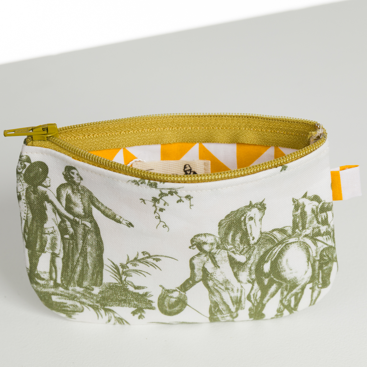 Le XCOMM_BIC BALLON PESTO est une petite pochette à fermeture éclair avec une fermeture éclair et une doublure jaunes, un tissu blanc avec des impressions pastorales vertes de personnes et de chevaux, et une étiquette latérale orange et blanche. la marque Toile de Jouy