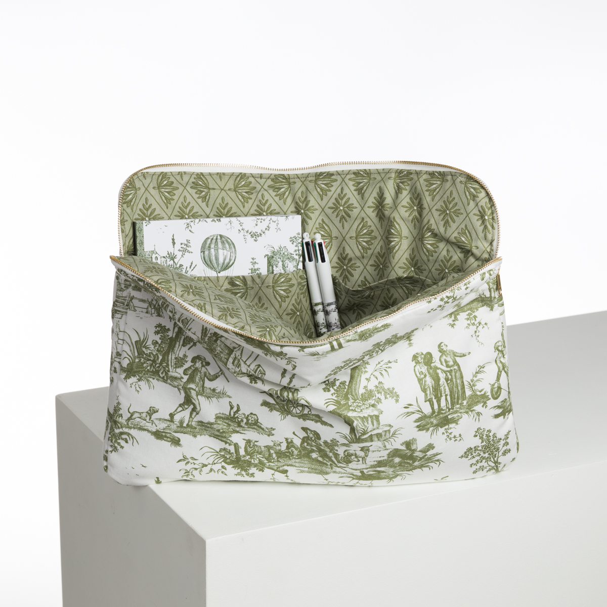 La pochette en tissu XCOMM_BIC BALLON PESTO, avec un motif toile vert et blanc et une doublure intérieure à motifs, est ouverte sur une surface blanche avec un carnet et deux stylos visibles à l'intérieur. la marque Toile de Jouy