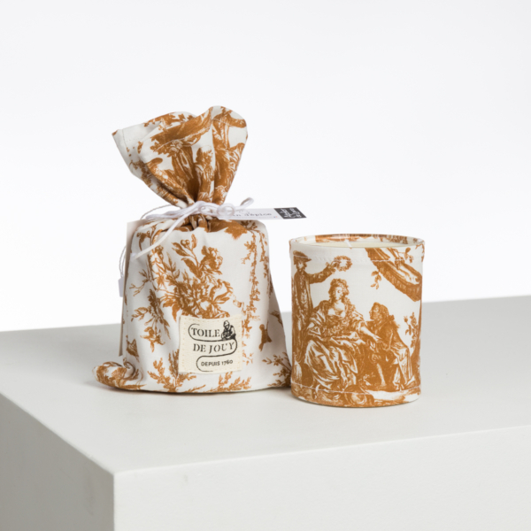 La bougie XCOMM_BIC BALLON PESTO, enveloppée dans un élégant tissu blanc et or au motif de toile classique, est présentée à côté de sa pochette à cordon assortie sur une surface blanche. la marque Toile de Jouy