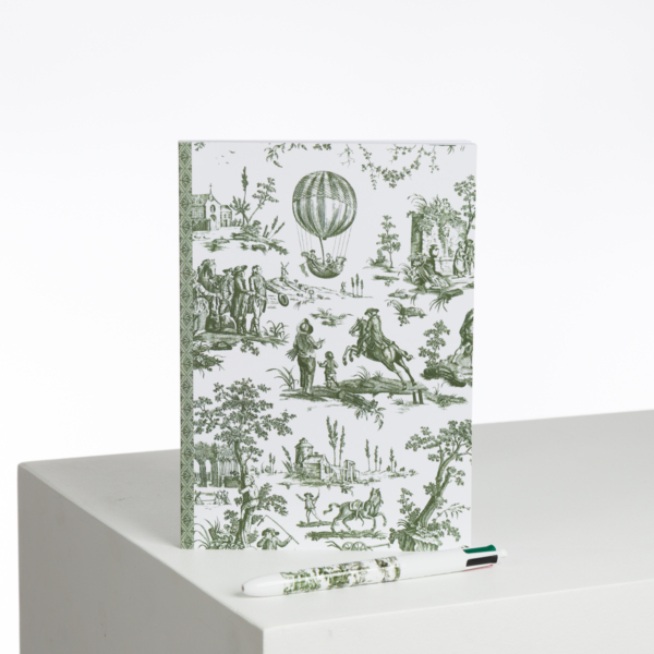 Le XCOMM_CARNET BROCHE A5 BALLON PESTO est un carnet blanc orné d'illustrations de ballons et de personnages de style vintage vert. Il se tient debout sur une surface blanche et est précédé d'un stylo assorti portant le même motif vert. la marque Toile de Jouy