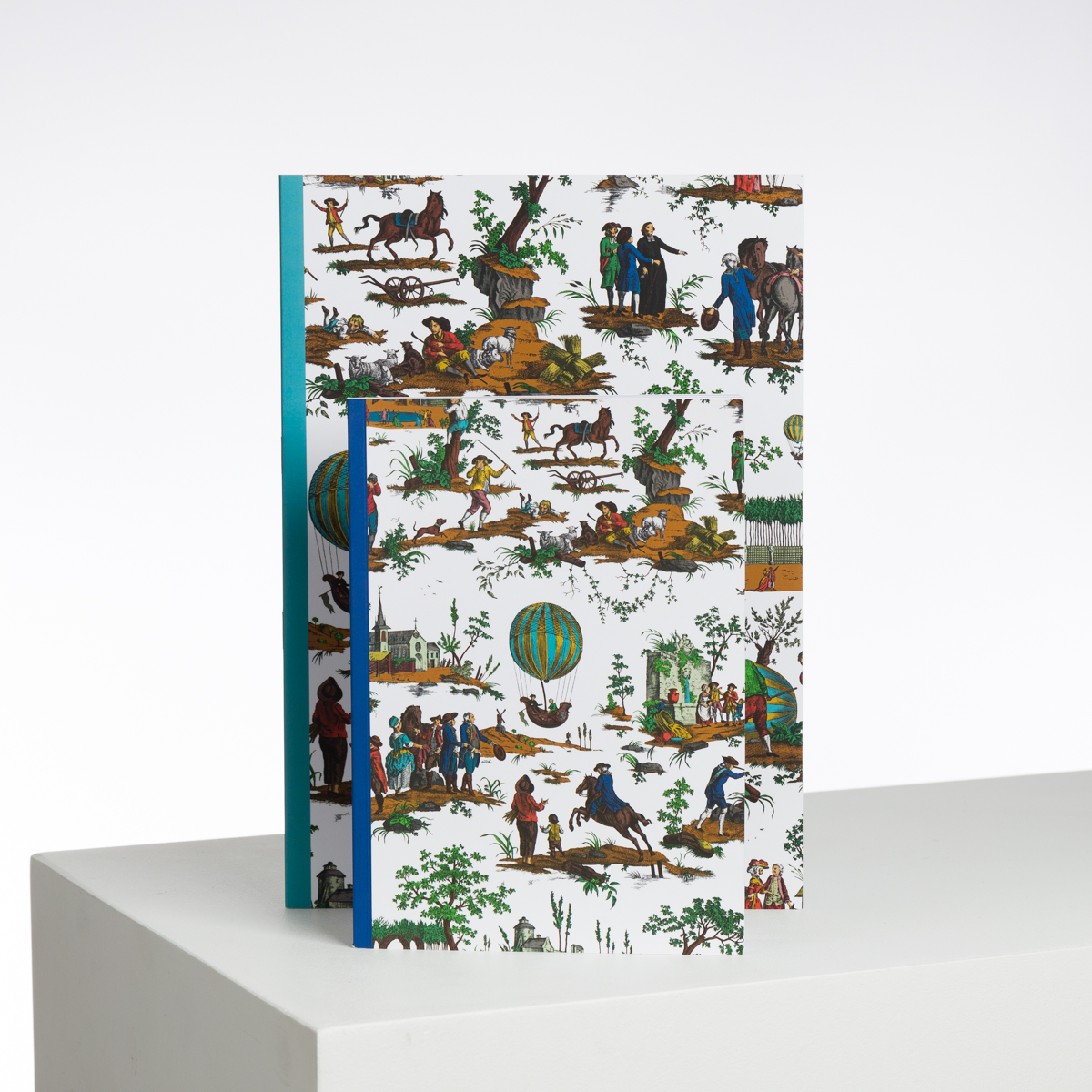 Deux carnets XCOMM_CAHIER A4 BALLON COL BD à couverture blanche présentent des scènes vibrantes de personnes, d'animaux et de montgolfières dans la nature, à l'endroit sur une surface blanche sur un fond uni. la marque Toile de Jouy