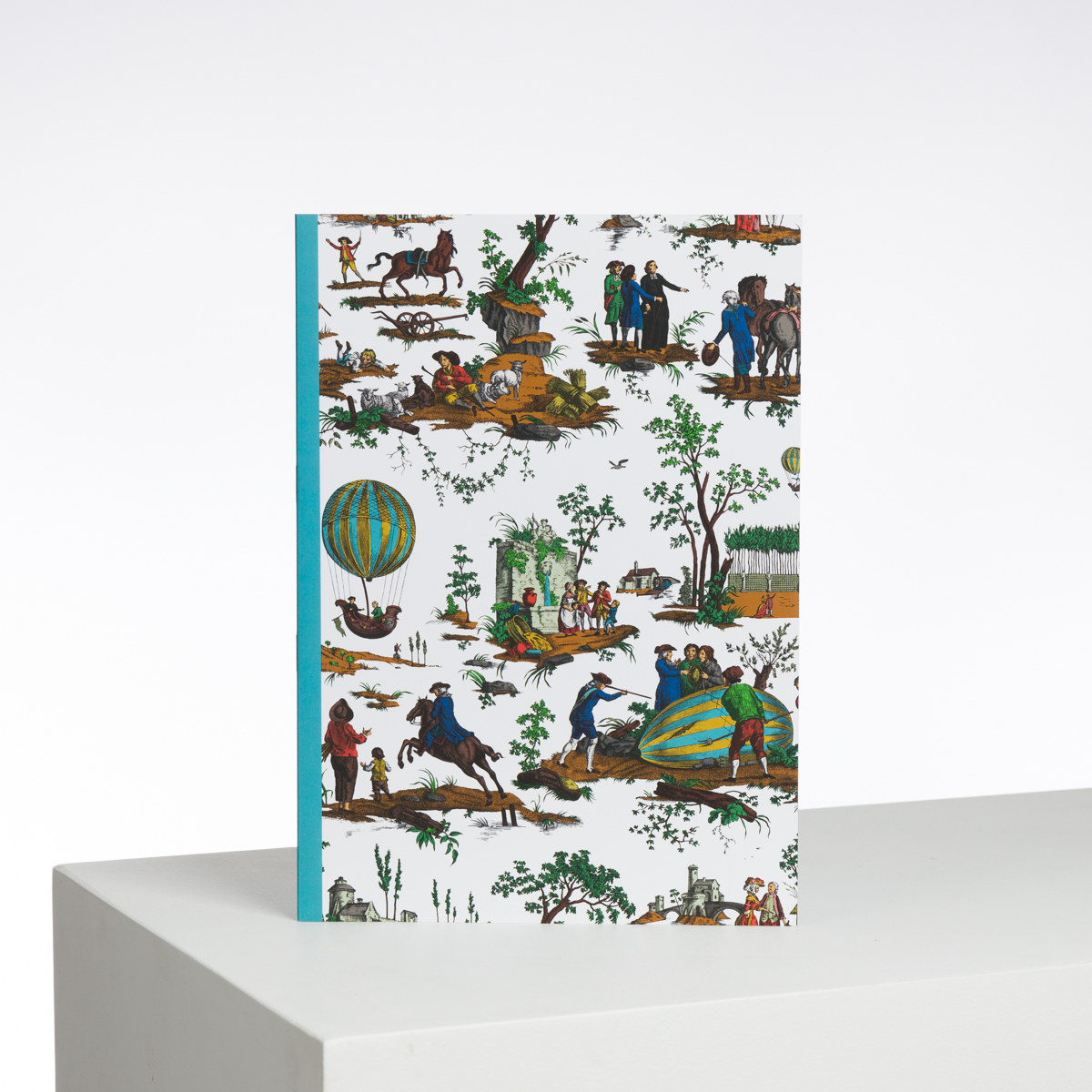 Le XCOMM_CAHIER A4 BALLON COL BD est un carnet dont la couverture colorée de style vintage présente des illustrations de personnes, d'animaux, de montgolfières et de scènes rurales, à l'endroit sur un fond blanc uni. la marque Toile de Jouy