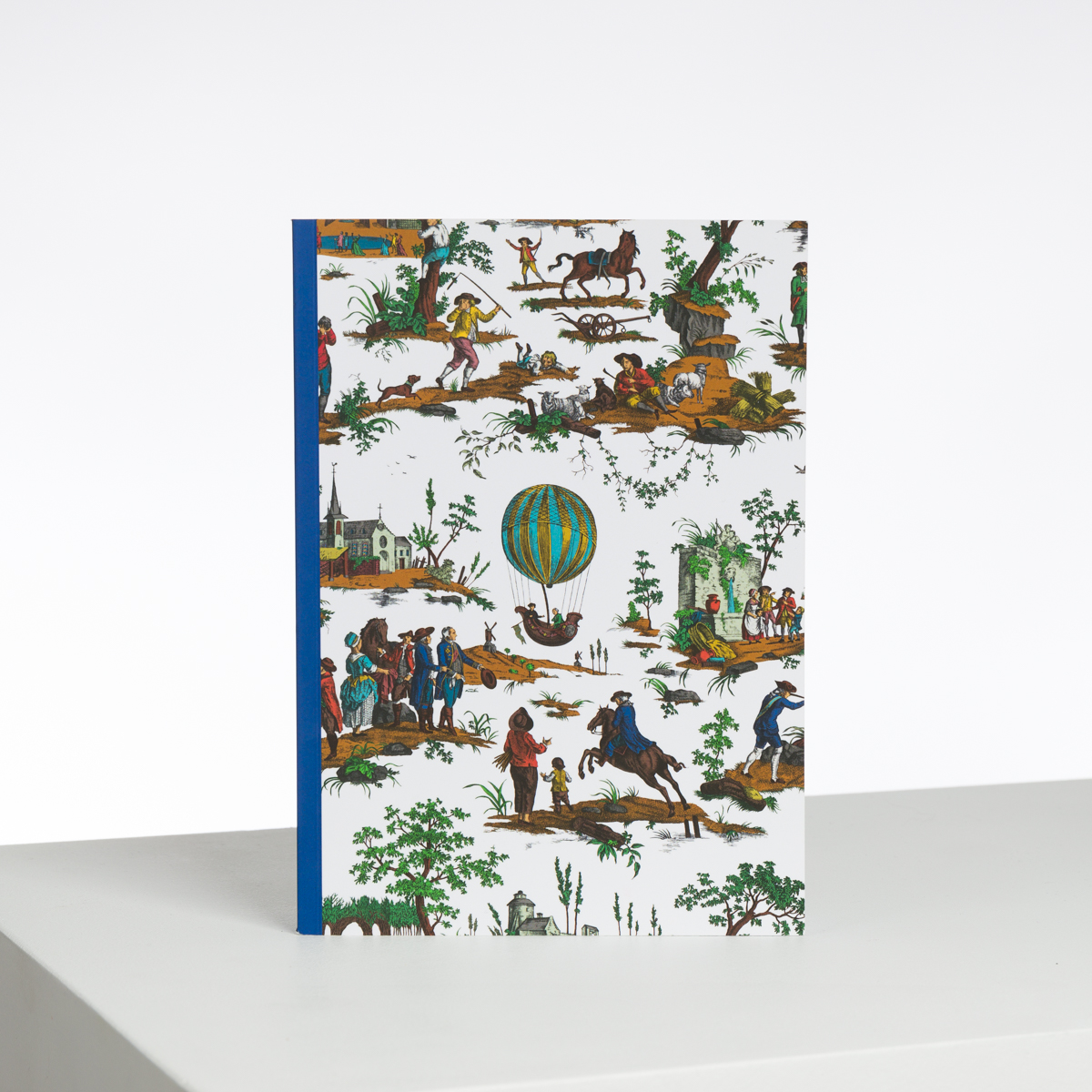 Le XCOMM_CAHIER A4 BALLON COL BD est un carnet à dos bleu dont la couverture présente des illustrations colorées de personnes, d'animaux, de montgolfières et de scènes extérieures, représentées debout sur une surface blanche sur fond uni. la marque Toile de Jouy