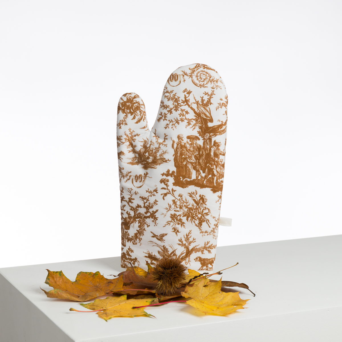Le XCOMM_BIC BALLON PESTO est un gant de cuisine blanc avec un motif de toile marron, debout sur une surface blanche avec des feuilles d'automne et une cosse de châtaigne. Le fond est uni et lumineux. la marque Toile de Jouy