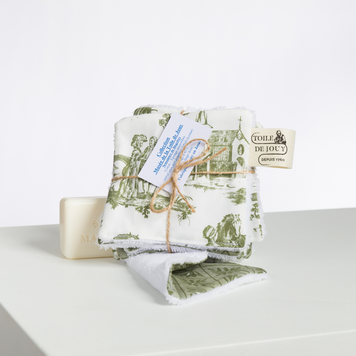 Le XCOMM_BIC BALLON PESTO présente des carrés de tissu blanc à motifs de toile, attachés avec de la ficelle et des étiquettes, représentant des scènes pastorales vintage, à côté d'une barre de savon de couleur claire sur une surface blanche. la marque Toile de Jouy