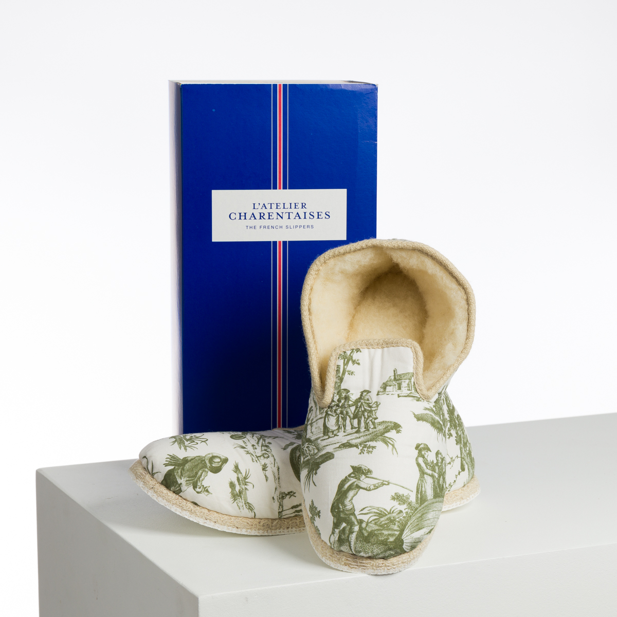 Une paire de pantoufles de couleur crème avec des illustrations pastorales vertes se trouve devant une boîte rectangulaire bleue étiquetée "XCOMM_BIC BALLON PESTO". la marque Toile de Jouy