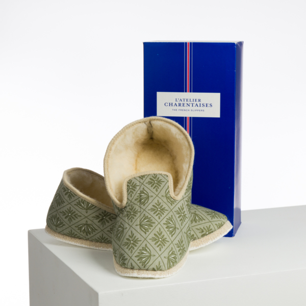 Une paire de chaussons XCOMM_BIC BALLON PESTO à motifs verts avec des intérieurs crème est présentée devant une grande boîte bleue "L'Atelier Charentaises The French Slippers", reposant sur une surface blanche. la marque Toile de Jouy