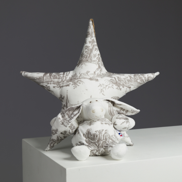 Un lapin en peluche et une étoile, tous deux en tissu blanc à motifs de toile grise, sont posés sur un piédestal blanc sur fond gris. la marque Toile de Jouy