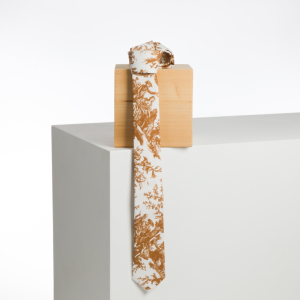 La XCOMM_BIC BALLON PESTO, une cravate blanche ornée d'un motif floral marron complexe, est présentée drapée sur un bloc de bois clair, sur une surface blanche minimaliste à fond uni. la marque Toile de Jouy