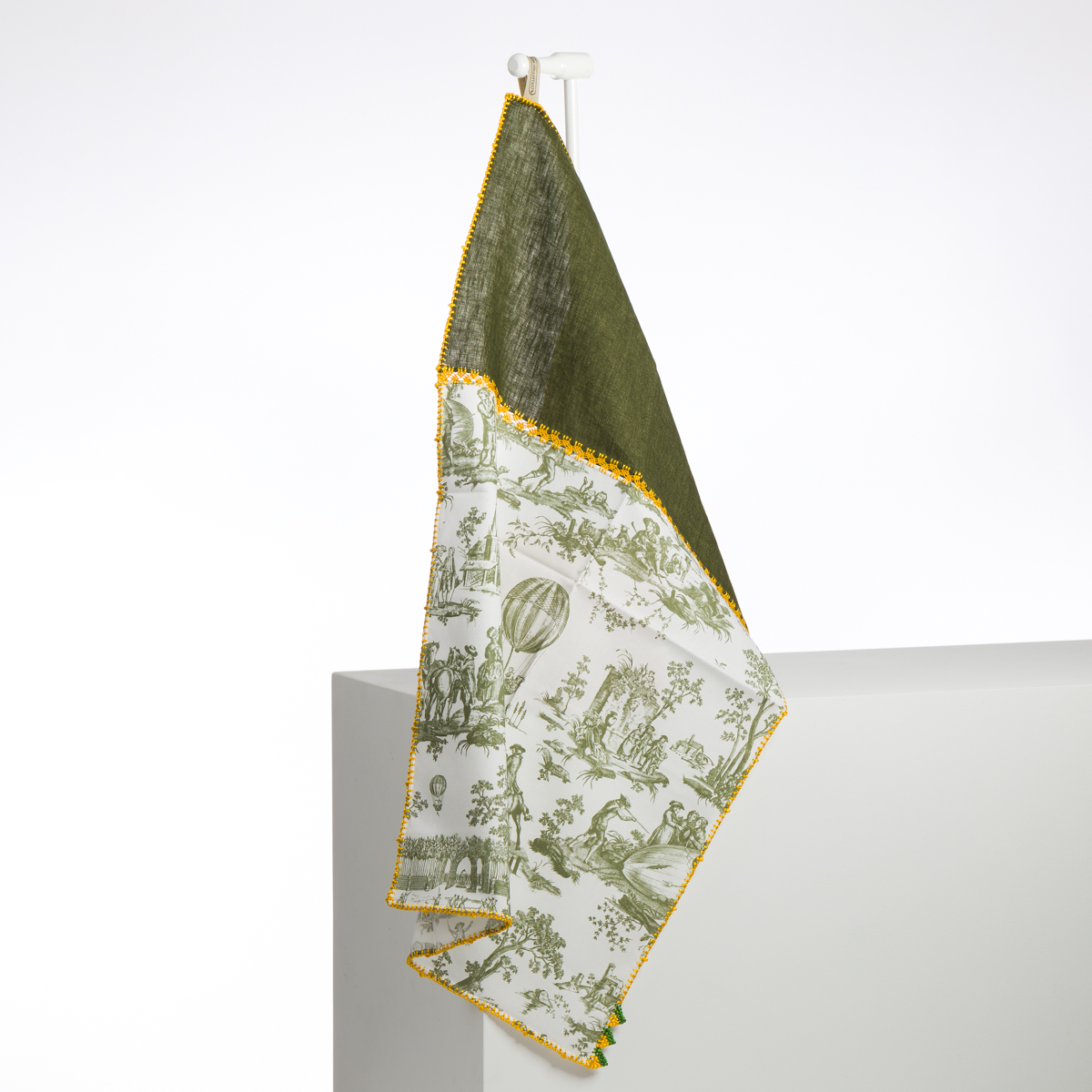 La serviette XCOMM_GRAND TORCHON BRODE ROSIERE CUMIN est suspendue à un crochet blanc et présente une séparation en diagonale : un côté vert olive uni, l'autre avec des motifs de toile blancs et verts, le tout bordé d'un liseré jaune. la marque Toile de Jouy