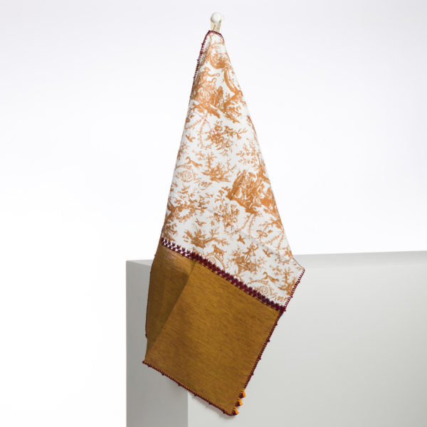 Le XCOMM_GRAND TORCHON BRODE ROSIERE CUMIN présente un motif de toile marron et blanc sur la partie supérieure et une partie inférieure jaune moutarde unie, suspendue à un crochet sur un fond blanc. la marque Toile de Jouy