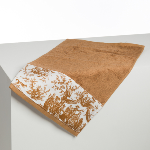 La serviette XCOMM_BIC BALLON PESTO est une serviette brune avec une bande blanche décorative représentant une scène pastorale brune complexe, drapée sur le bord d'une surface blanche. la marque Toile de Jouy