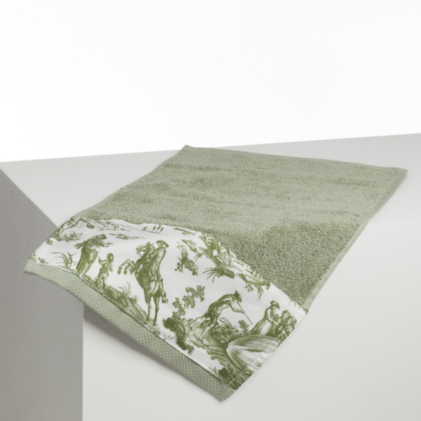 La serviette XCOMM_BIC BALLON PESTO est une serviette de bain vert clair avec une bordure blanche présentant des motifs pastoraux et naturels verts, élégamment drapés sur le bord d'une surface blanche. la marque Toile de Jouy