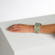 Le bras d'une personne repose sur une surface blanche. Elle porte le XCOMM_BIC BALLON PESTO, un large bracelet vert clair avec un motif de feuilles. L'arrière-plan est uni et blanc. la marque Toile de Jouy