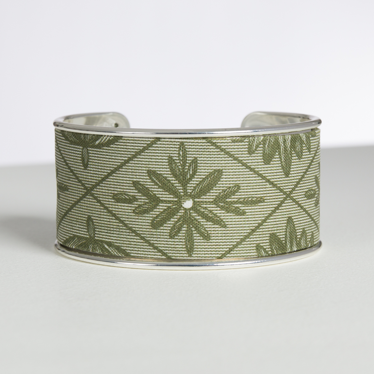 Le XCOMM_BIC BALLON PESTO est un large bracelet manchette métallique avec un motif géométrique vert olive composé de formes étoilées et de lignes diagonales sur un fond de tissu texturé. la marque Toile de Jouy