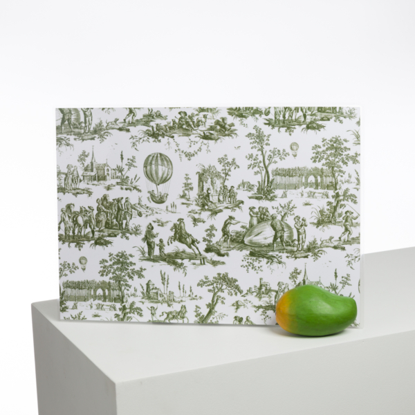 Le panneau XCOMM_BIC BALLON PESTO présente des illustrations pastorales vertes de personnes, d'arbres et de montgolfières, debout sur une surface blanche. Une mangue mûre verte et jaune est posée devant le panneau. la marque Toile de Jouy