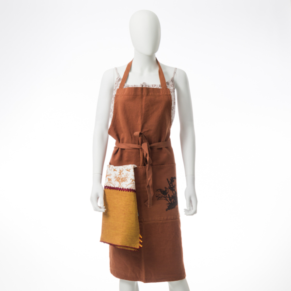 Un mannequin blanc porte le XCOMM_GRAND TORCHON BRODE ROSIERE CUMIN - un tablier de couleur rouille avec une poche sur le devant, une serviette en tissu attachée et des coutures décoratives - sur un fond blanc uni. la marque Toile de Jouy