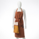 Un mannequin blanc porte le XCOMM_GRAND TORCHON BRODE ROSIERE CUMIN - un tablier de couleur rouille avec une poche sur le devant, une serviette en tissu attachée et des coutures décoratives - sur un fond blanc uni. la marque Toile de Jouy