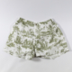 Le short XCOMM_BIC BALLON PESTO présente un imprimé de toile pastorale verte avec des personnages, des arbres et des scènes rurales sur un tissu blanc, une ceinture élastique et est présenté à plat sur un fond blanc uni. la marque Toile de Jouy