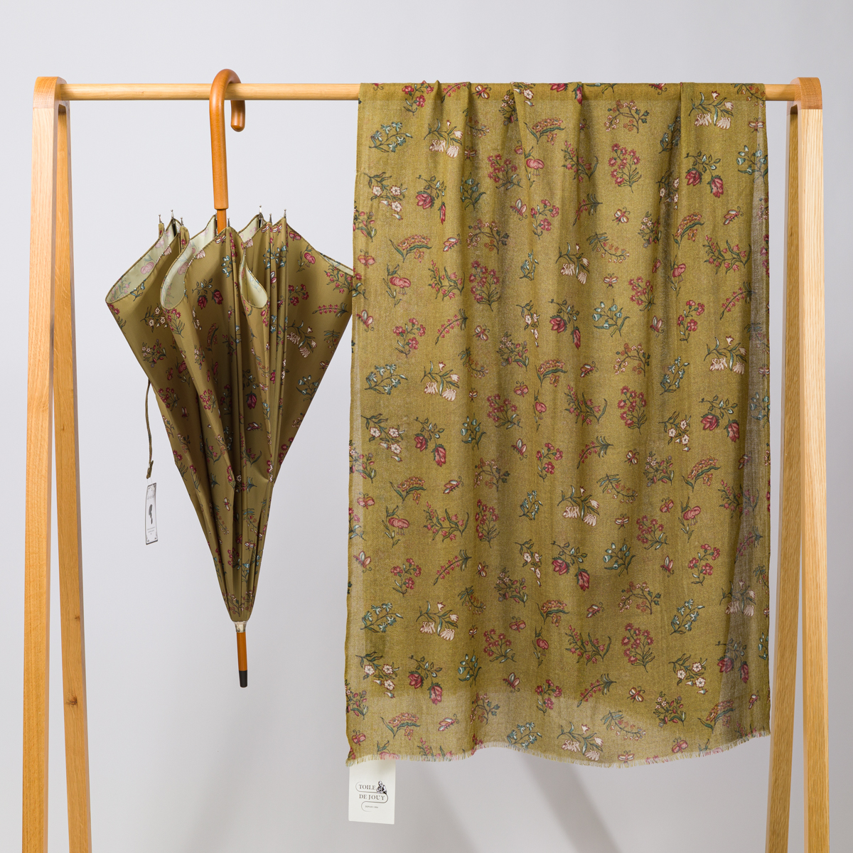 Un parapluie vert « FANTAISIE INDIENNE » de la marque ETIQUETTE DE VOYAGE et une écharpe assortie, ornés d’un motif floral et de lapins, sont suspendus à un porte-parapluies en bois sur un fond clair uni. la marque Toile de Jouy