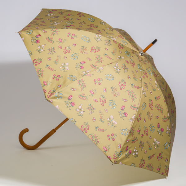 Le parapluie « FANTAISIE INDIENNE » de la marque ETIQUETTE DE VOYAGE, doté d’une toile jaune ornée d’un motif de petites fleurs colorées et d’un manche incurvé en bois, est ouvert et posé sur une surface blanche. la marque Toile de Jouy