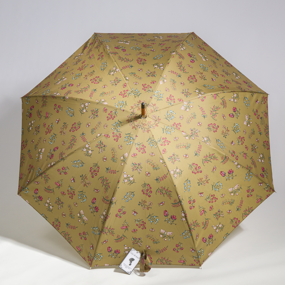 Un parapluie jaune entièrement ouvert de la marque ETIQUETTE DE VOYAGE « FANTAISIE INDIENNE », orné d'un motif de petites fleurs colorées et d'abeilles. Une étiquette est fixée à la poignée, près du bord inférieur. Le fond est blanc uni. la marque Toile de Jouy