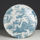 Le XCOMM_PLATEAU ROND 46CM DELICES BLEU est une assiette décorative ronde représentant des scènes pastorales bleues avec des personnes, des animaux et des arbres sur un fond blanc. Elle est présentée debout sur un support blanc sur un fond sombre. la marque Toile de Jouy