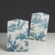 Le XCOMM_PLATEAU ROND 46CM DELICES BLEU, représentant des scènes pastorales bleues sur fond blanc, se tient debout sur un support blanc sur un fond sombre et une surface claire. la marque Toile de Jouy