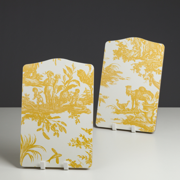 Le XCOMM_PLATEAU ROND 46CM DELICES BLEU présente des scènes pastorales d'un jaune éclatant avec des personnes, des arbres et des animaux sur un fond blanc, présentées sur des supports blancs sur une toile de fond neutre et sombre. la marque Toile de Jouy