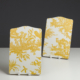 Le XCOMM_PLATEAU ROND 46CM DELICES BLEU présente des scènes pastorales d'un jaune éclatant avec des personnes, des arbres et des animaux sur un fond blanc, présentées sur des supports blancs sur une toile de fond neutre et sombre. la marque Toile de Jouy