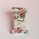 Un savon enveloppé dans du papier blanc orné d’un motif floral coloré. L’étiquette porte la mention « ETIQUETTE DE VOYAGE ‘FANTAISIE INDIENNE’ ». Le savon est représenté sur un fond rose pâle et blanc. la marque Toile de Jouy
