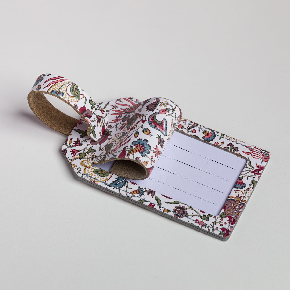 Une étiquette à bagages « FANTAISIE INDIENNE » de la marque ETIQUETTE DE VOYAGE, ornée d’un motif floral aux couleurs vives, est présentée avec son rabat partiellement ouvert, laissant apparaître une fiche nominative vierge à l’intérieur. La lanière, confectionnée dans le même tissu à motifs, est fixée en boucle. L’étiquette est mise en valeur sur un fond blanc uni. la marque Toile de Jouy