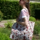 Une femme portant des lunettes de soleil et un t-shirt rayé se tient à l'extérieur, tenant dans ses mains le sac fourre-tout ETIQUETTE DE VOYAGE « FANTAISIE INDIENNE » ; on aperçoit de la verdure et des fleurs en arrière-plan tandis qu'elle est tournée vers la gauche. la marque Toile de Jouy