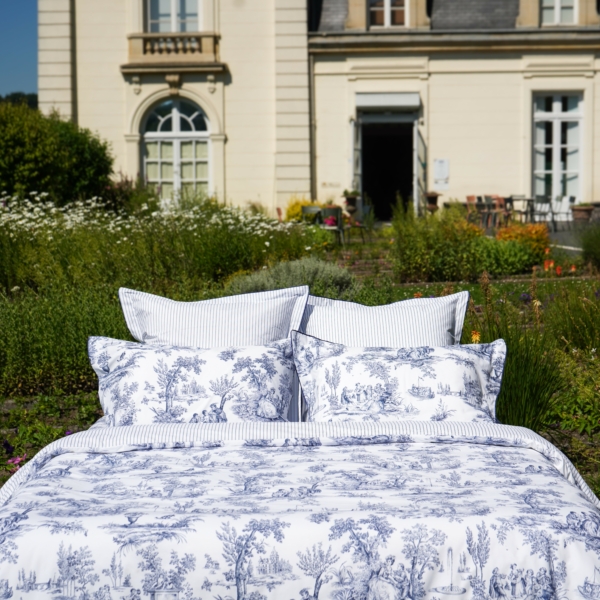 La literie XCOMM_PEIGNOIR M ou L en toile blanche et bleue est présentée sur un lit à l'extérieur, dans un jardin, devant une élégante bâtisse de couleur crème avec de grandes fenêtres et des portes cintrées. la marque Toile de Jouy