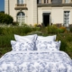 La literie XCOMM_PEIGNOIR M ou L en toile blanche et bleue est présentée sur un lit à l'extérieur, dans un jardin, devant une élégante bâtisse de couleur crème avec de grandes fenêtres et des portes cintrées. la marque Toile de Jouy