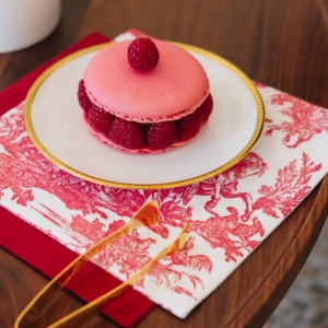 Sur une table en bois, un macaron rose avec des framboises fraîches repose sur une assiette blanche bordée d'or, à côté d'une serviette SERVIETTES 33X33 "LES 4 PARTIES DU MONDE" ROUGE GARANCE et d'une décoration en caramel. la marque Toile de Jouy
