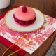 Sur une table en bois, un macaron rose avec des framboises fraîches repose sur une assiette blanche bordée d'or, à côté d'une serviette SERVIETTES 33X33 "LES 4 PARTIES DU MONDE" ROUGE GARANCE et d'une décoration en caramel. la marque Toile de Jouy
